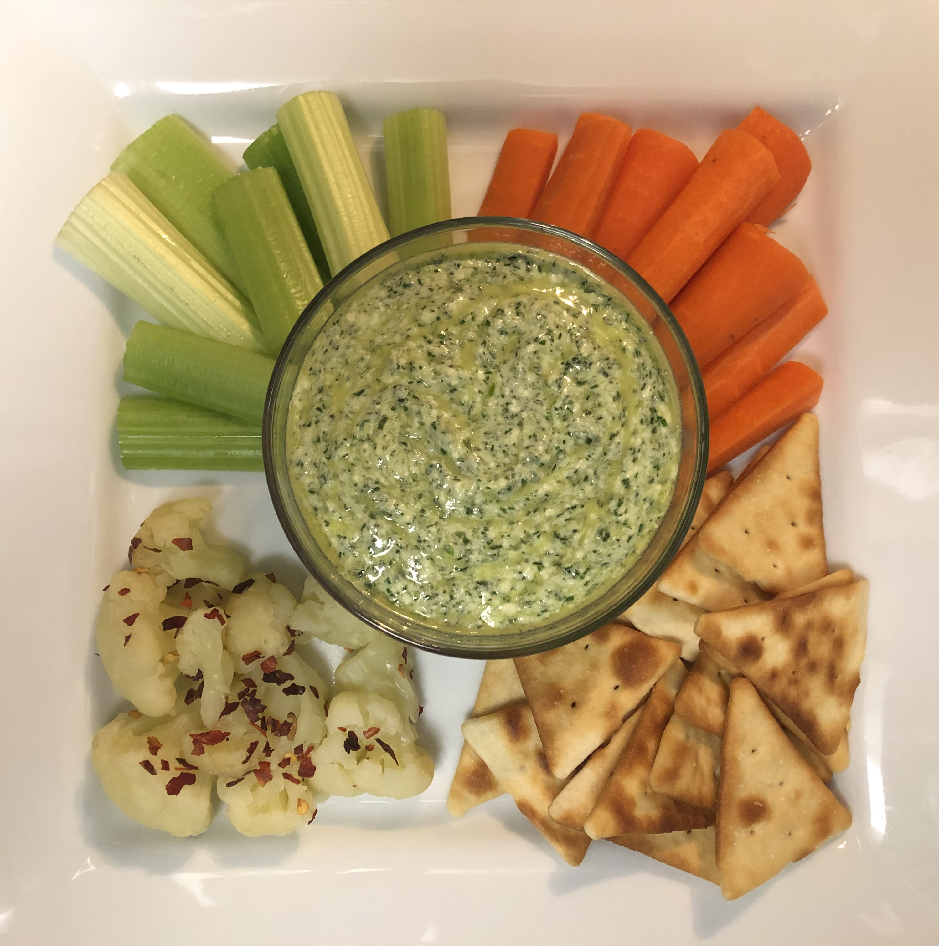 Green Goddess dip plated in a bowl in center of plate. Surrounding the bowl is carrots, celery, pita chips, and cauliflower with red pepper flakes.
