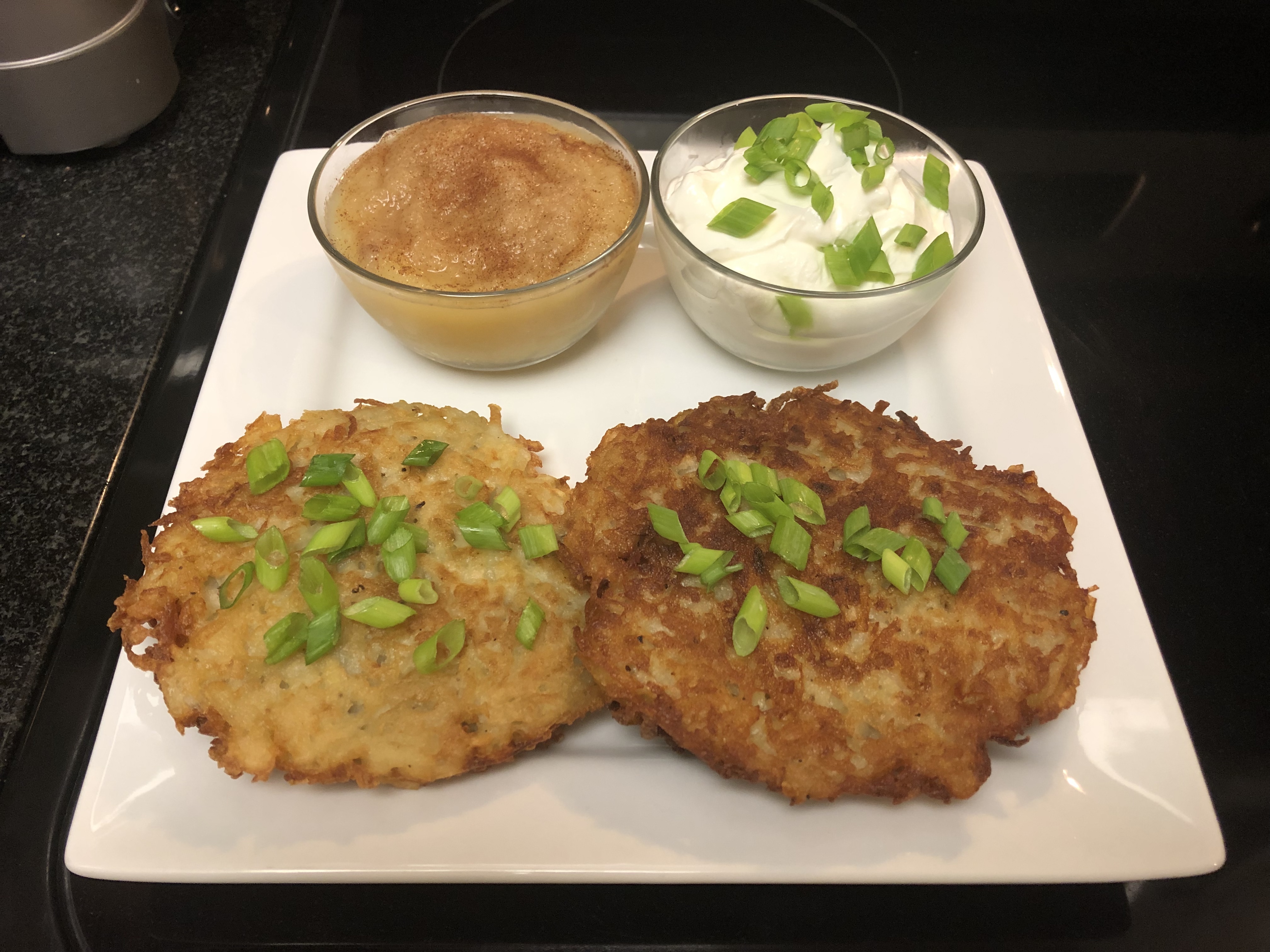 Potato pancakes plated and garnished with green onions with side bowls of garnished apple sauce and sour cream